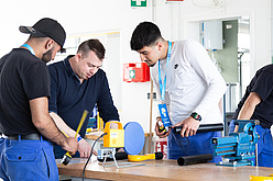 Fachmann instruiert zwei Lehrlinge an Tisch mit Maschinen und Werkzeugen